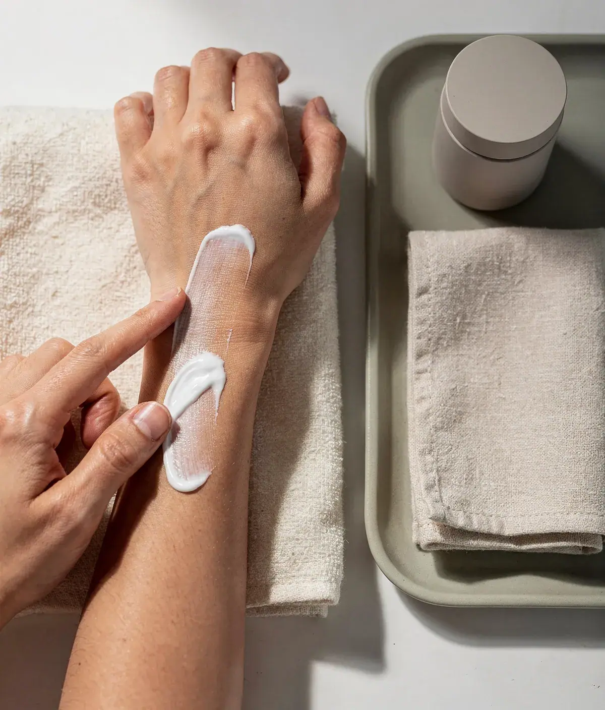 Antebrazo adulto con crema corporal, pañuelo de algodón y envase neutro en una escena editorial de absorción.