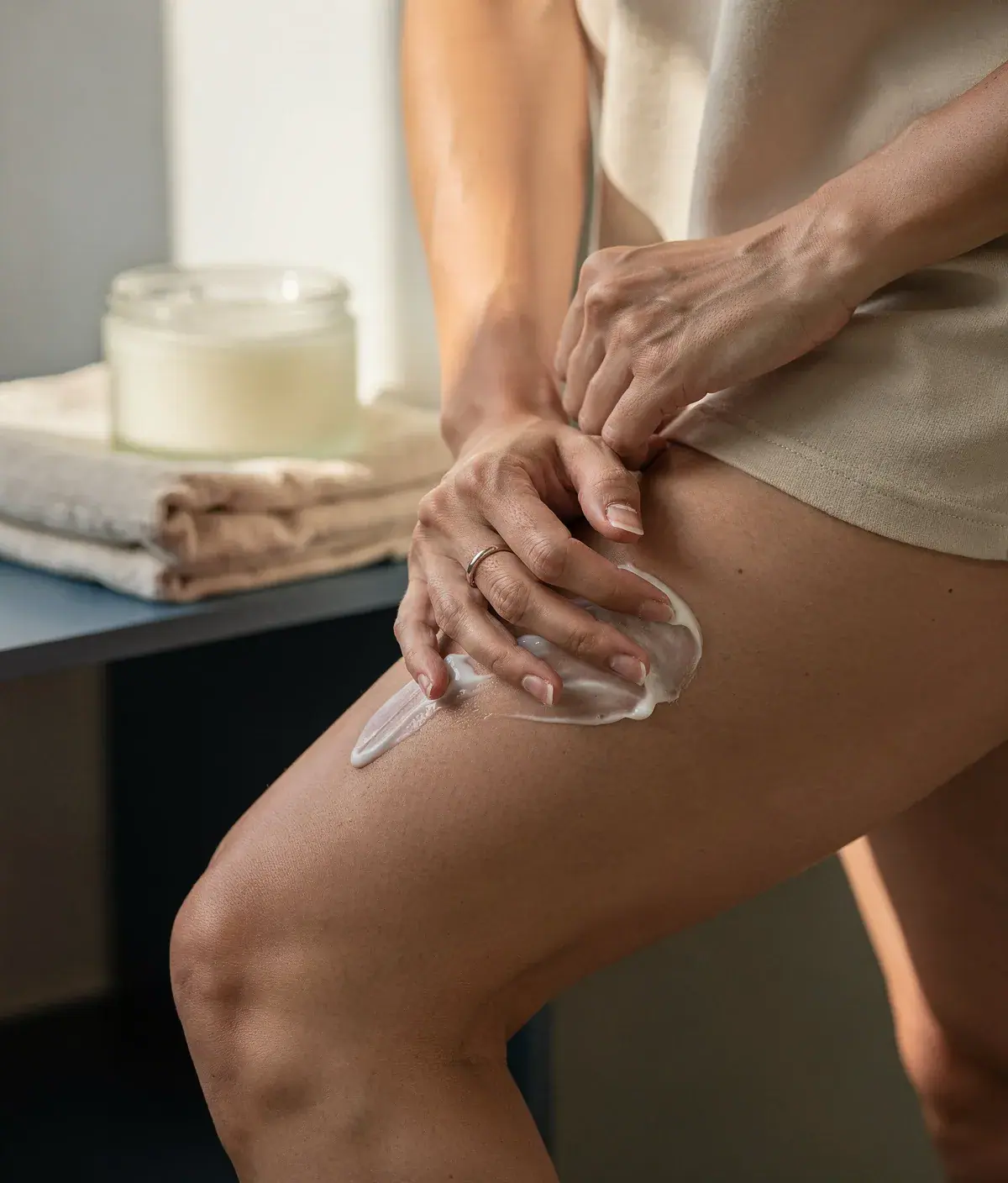 Mujer adulta masajeando el muslo con crema corporal junto a una toalla y un frasco.  